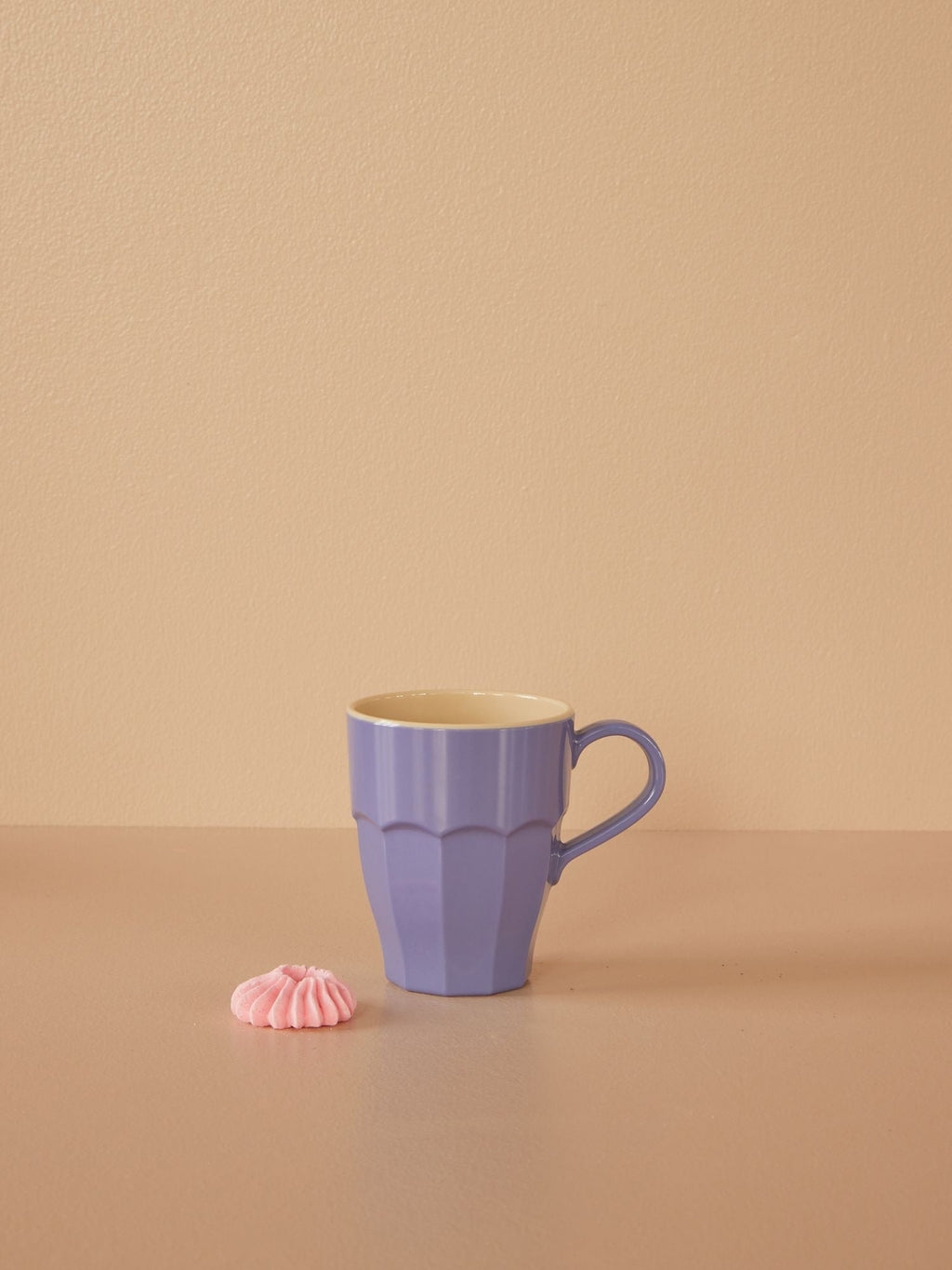 A lavender-colored melamine mug on a plain background, with a small pink object to the bottom right of the mug.